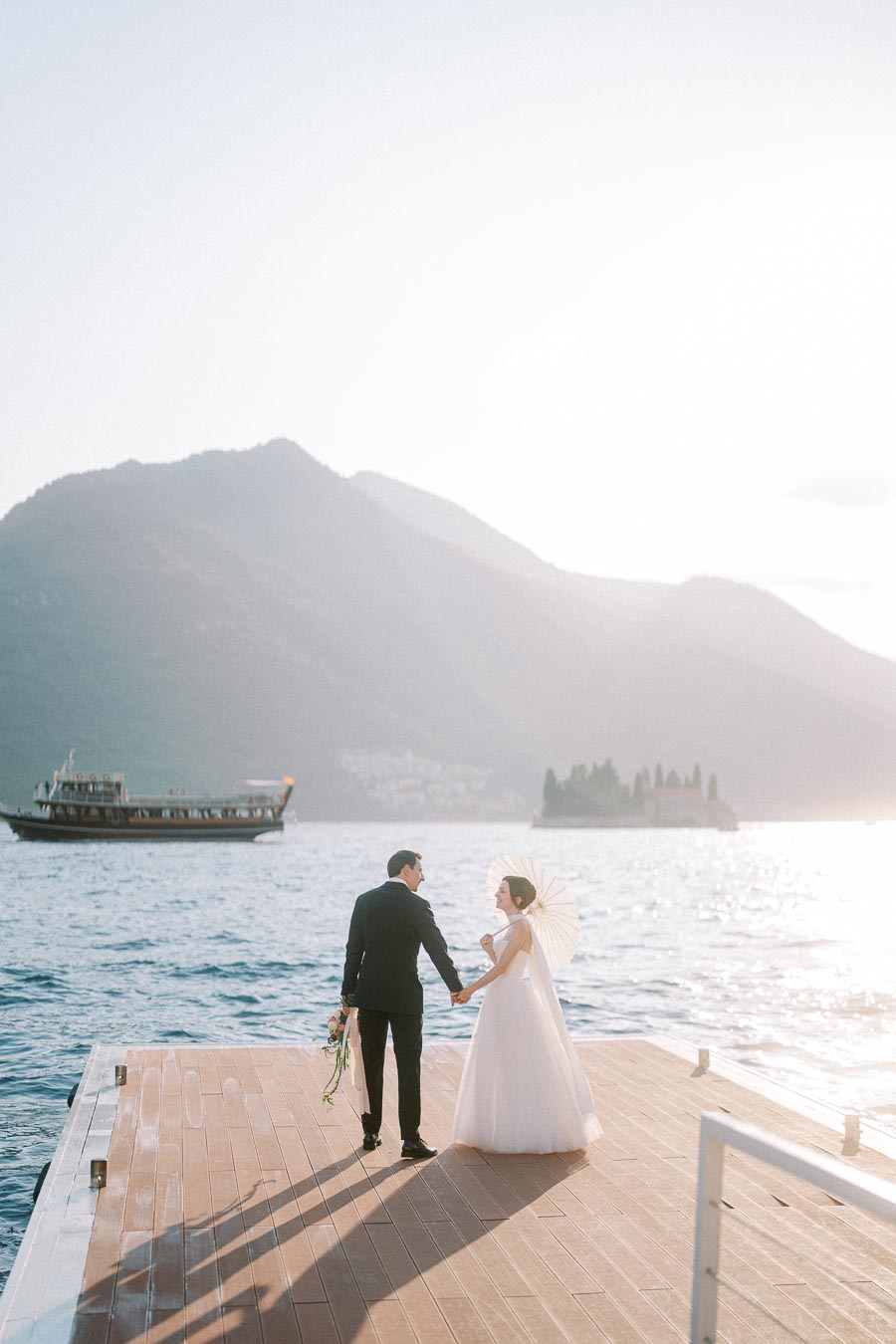 A bride and groom hold hands on a wooden dock by a scenic lake, with mountains in the background. The bride holds a white parasol and a bouquet of flowers, while a boat and a small island are visible on the water under the bright afternoon sun.