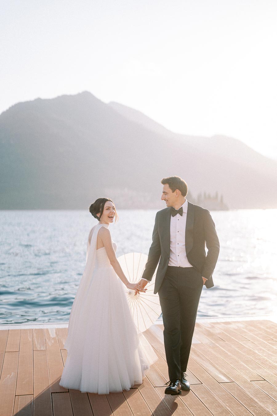 Wedding couple smiling and holding hands on a wooden pier with a scenic mountain and lake backdrop, bride wearing elegant white gown and groom in a classic black tuxedo.