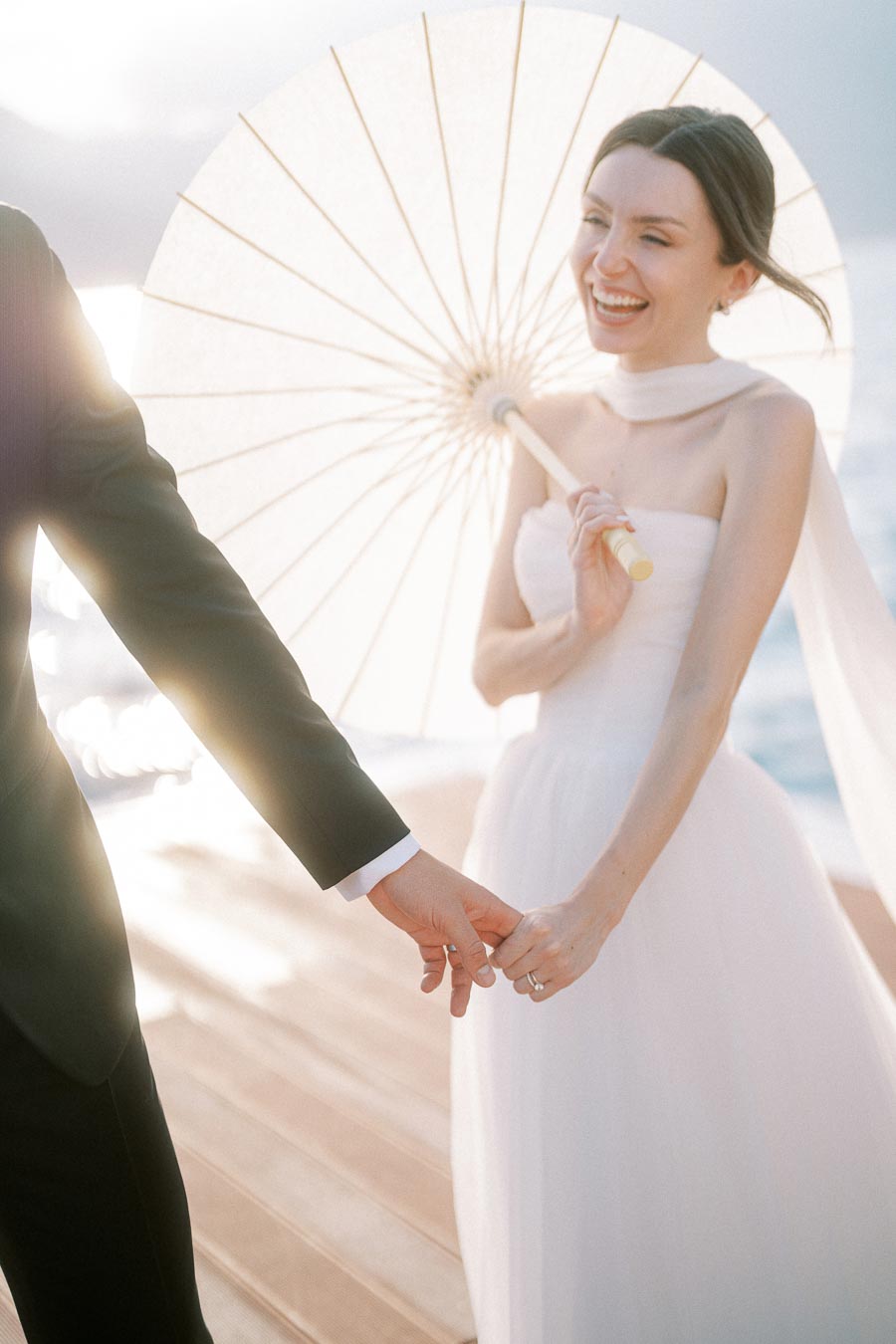 A bride holding a white parasol smiles joyfully while holding hands with her partner, dressed in a suit, on a sunny day by the water.