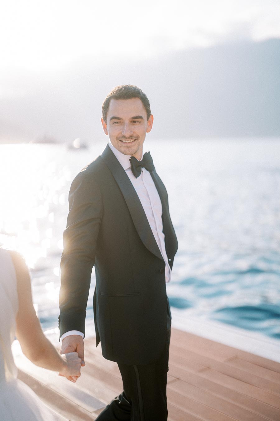 Groom in a tuxedo holding hands with the bride on a sunlit dock by the water, showcasing a romantic wedding moment.
