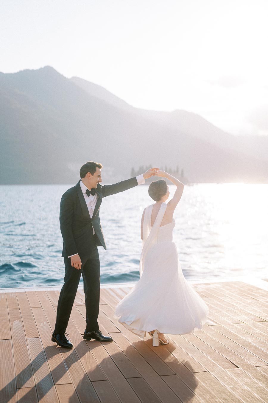 A bride and groom dance on a wooden deck by a serene lake, with mountains in the background, under a bright, sunlit sky.