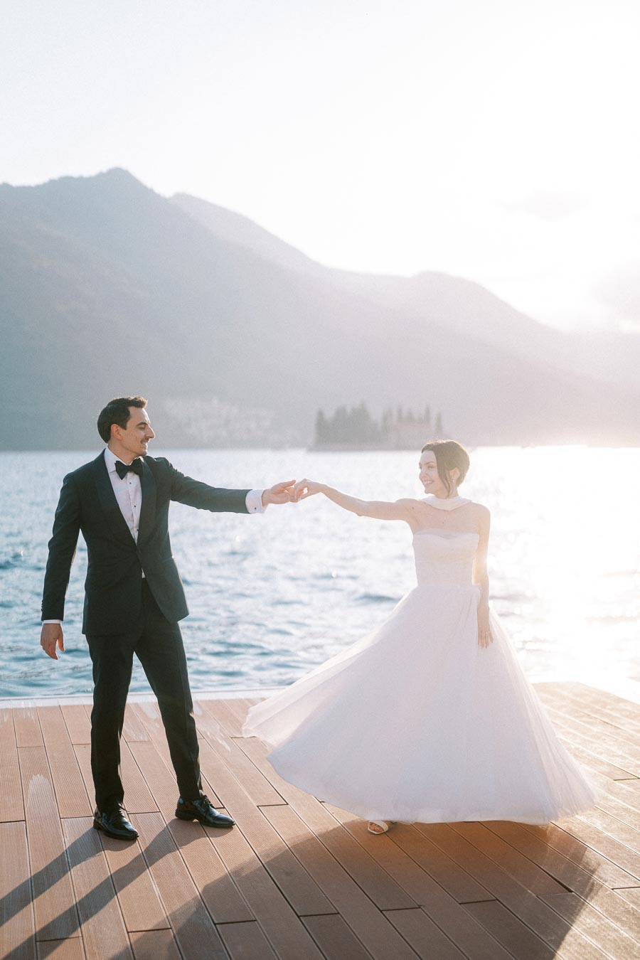 Bride and groom dancing on a wooden deck by a serene lake with mountains in the background, under warm sunlight.