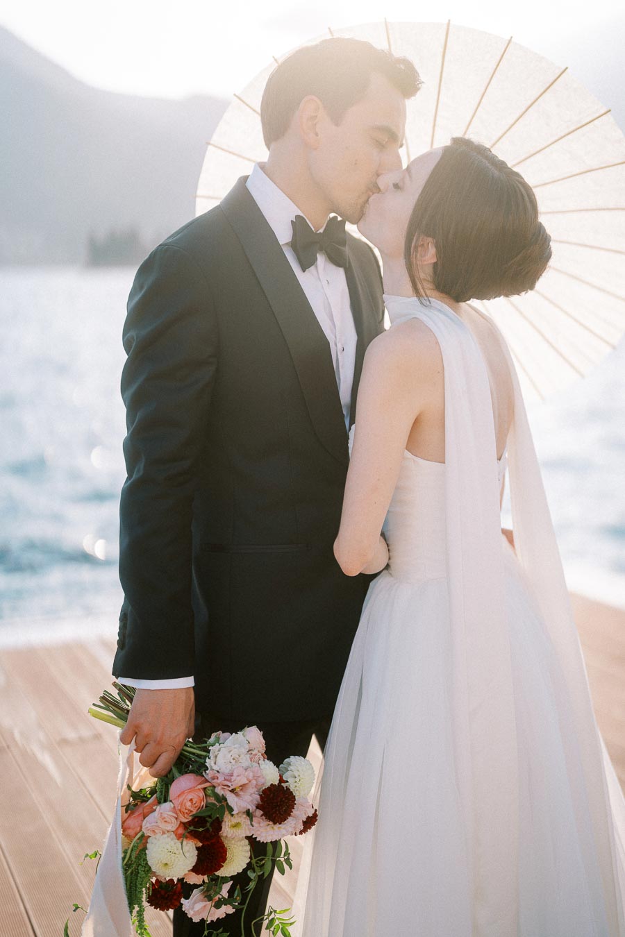 A bride and groom share a romantic kiss on a lakeside boardwalk, with the bride holding a bouquet of colorful flowers and a parasol providing a soft background glow. The groom is dressed in a formal black suit, perfect for a wedding celebration by the water.