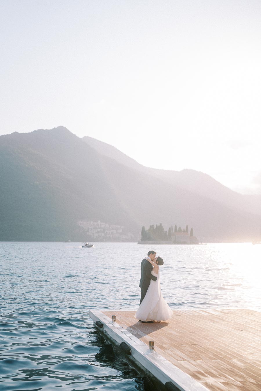 A romantic couple embraces on a wooden dock overlooking a picturesque lake, with a misty mountainous backdrop illuminated by sunlight.