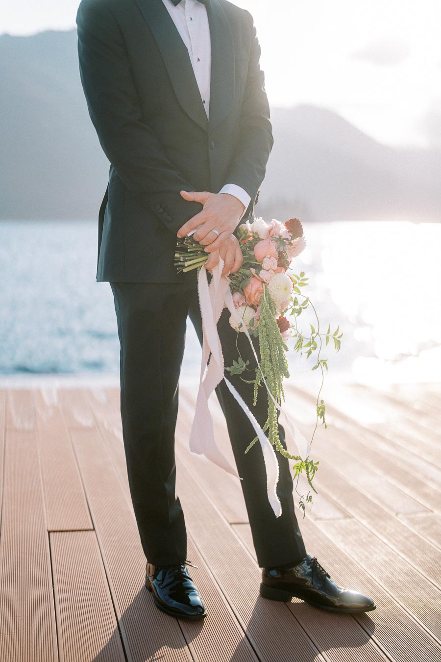 A groom in a black tuxedo holding a vibrant bouquet of pink and white flowers, standing on a wooden deck by the water with mountains in the background at sunset.