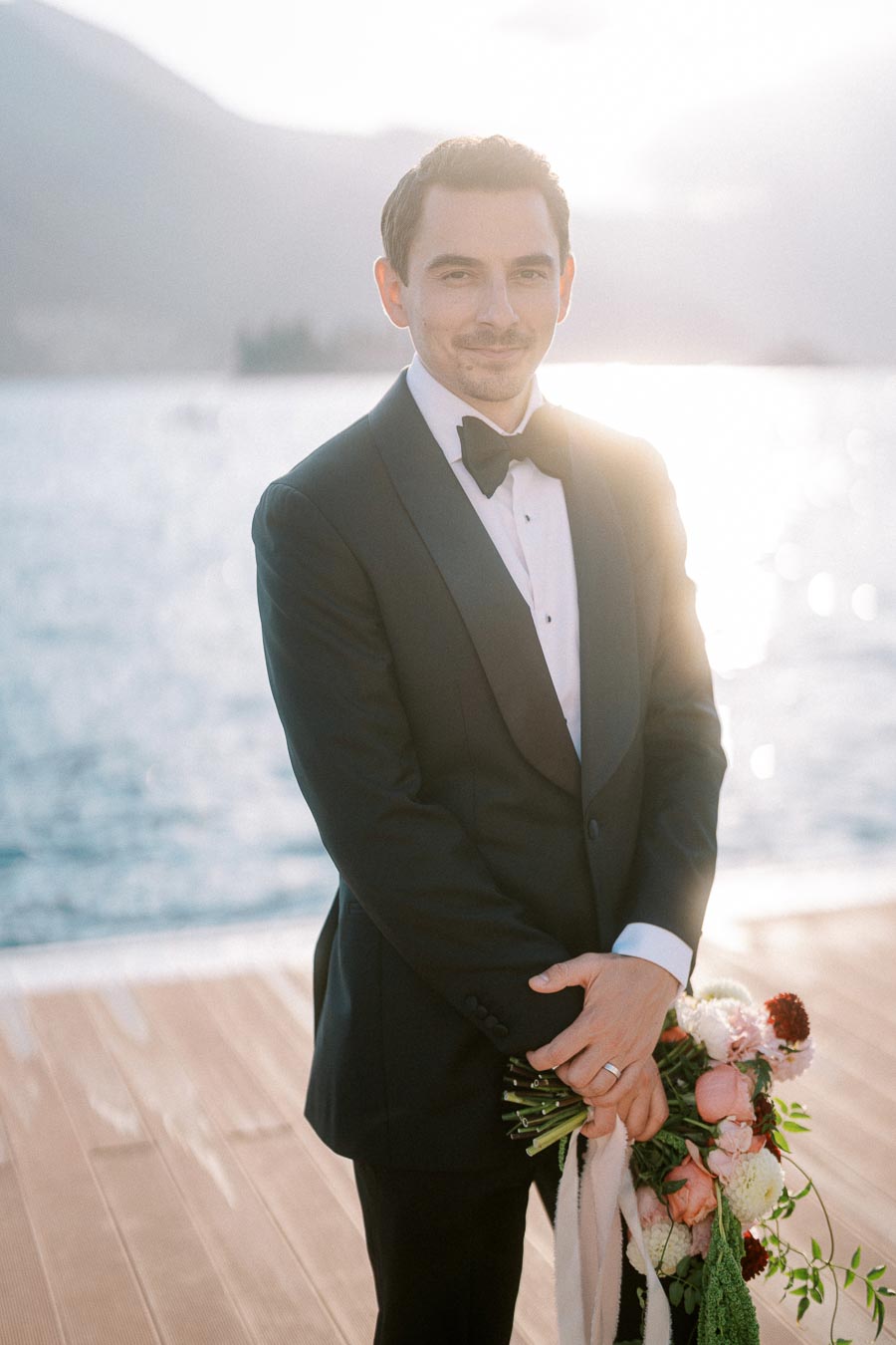Elegant groom in a tuxedo holding a bouquet, standing by a serene lakeside, with mountains and sunlight in the background.