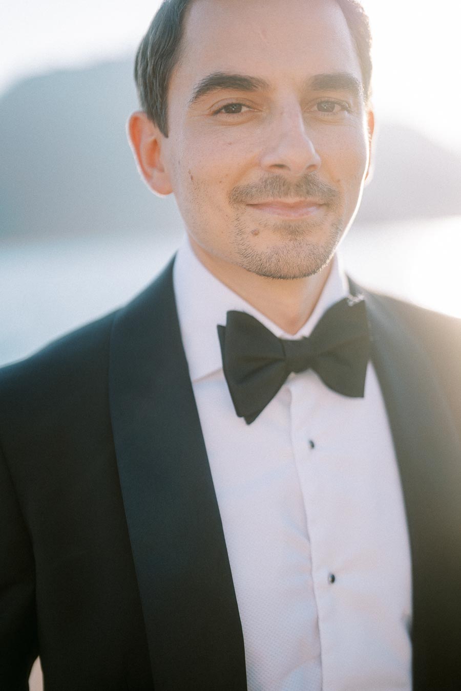 A man in a formal black tuxedo with a bow tie, standing outdoors with soft sunlight in the background.