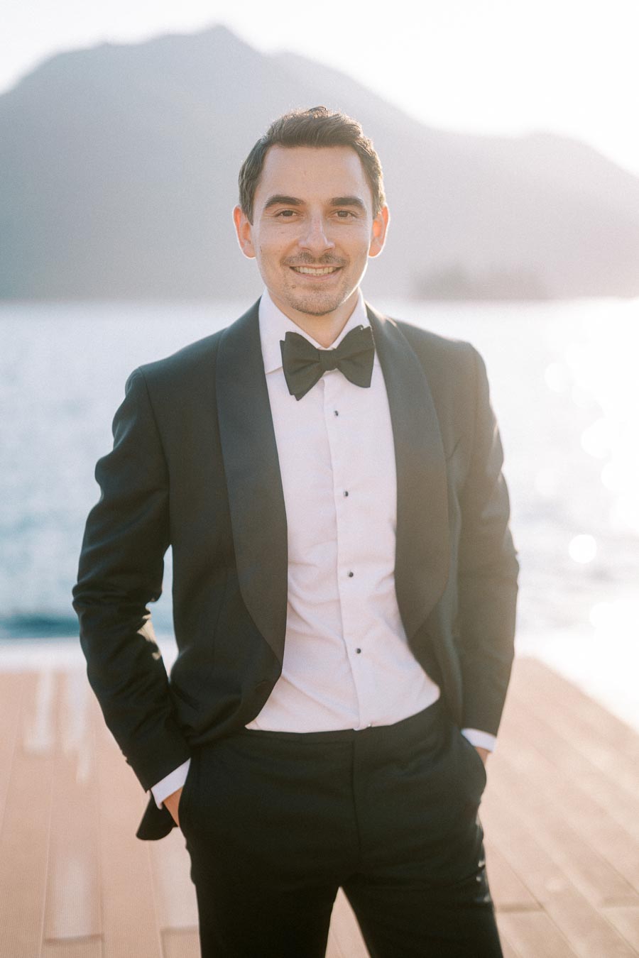 Man in a formal black tuxedo standing confidently on a wooden dock with a serene lake and mountains in the background, under soft natural light.