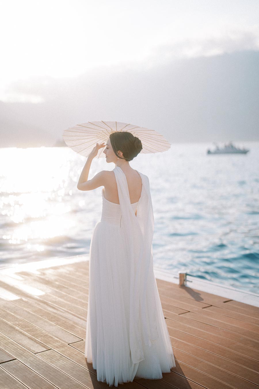 Elegant bride in flowing white gown holding parasol, standing on a wooden deck overlooking the serene ocean at sunset.