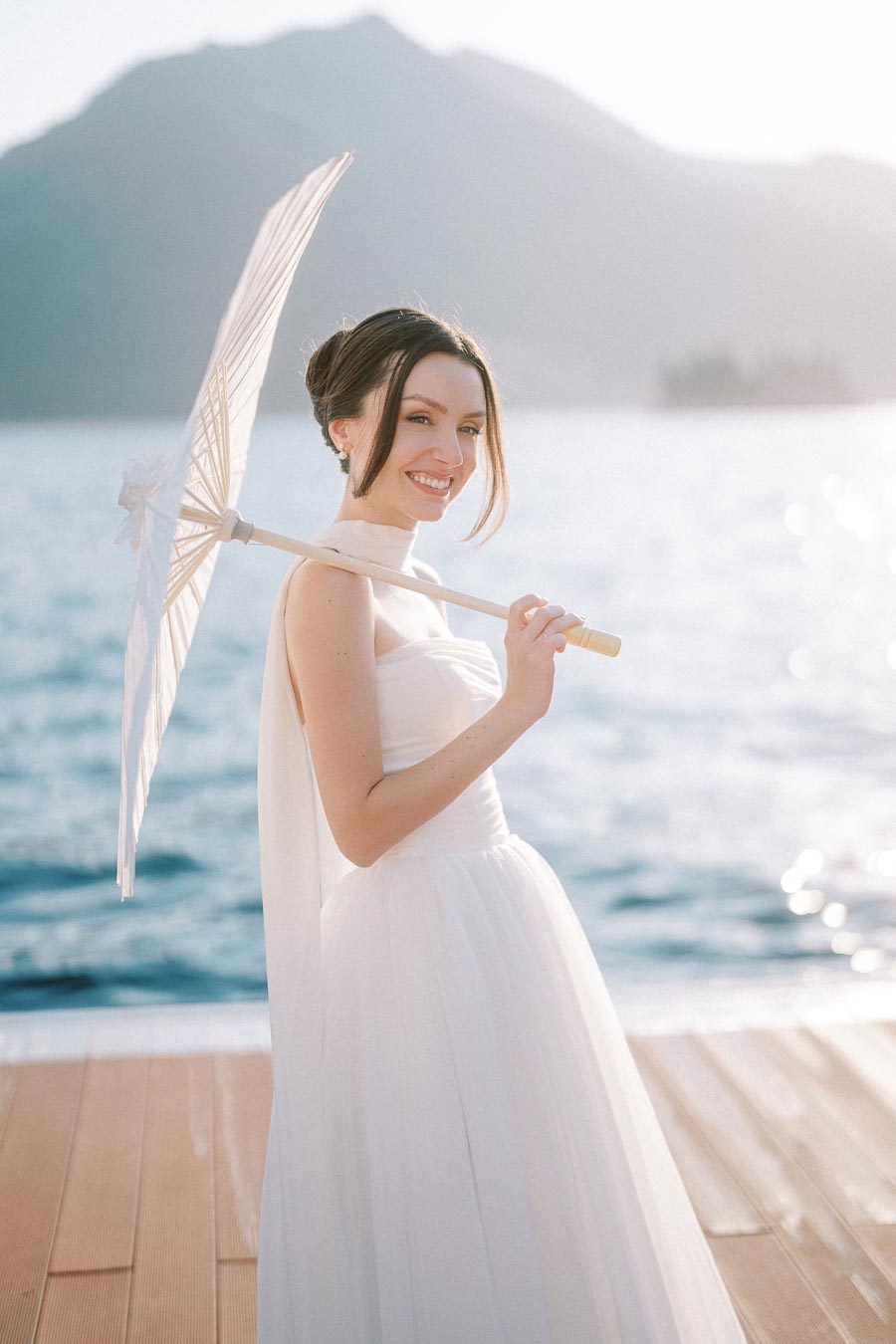 A woman in a white dress smiles while holding a parasol by a scenic lake with mountains in the background
