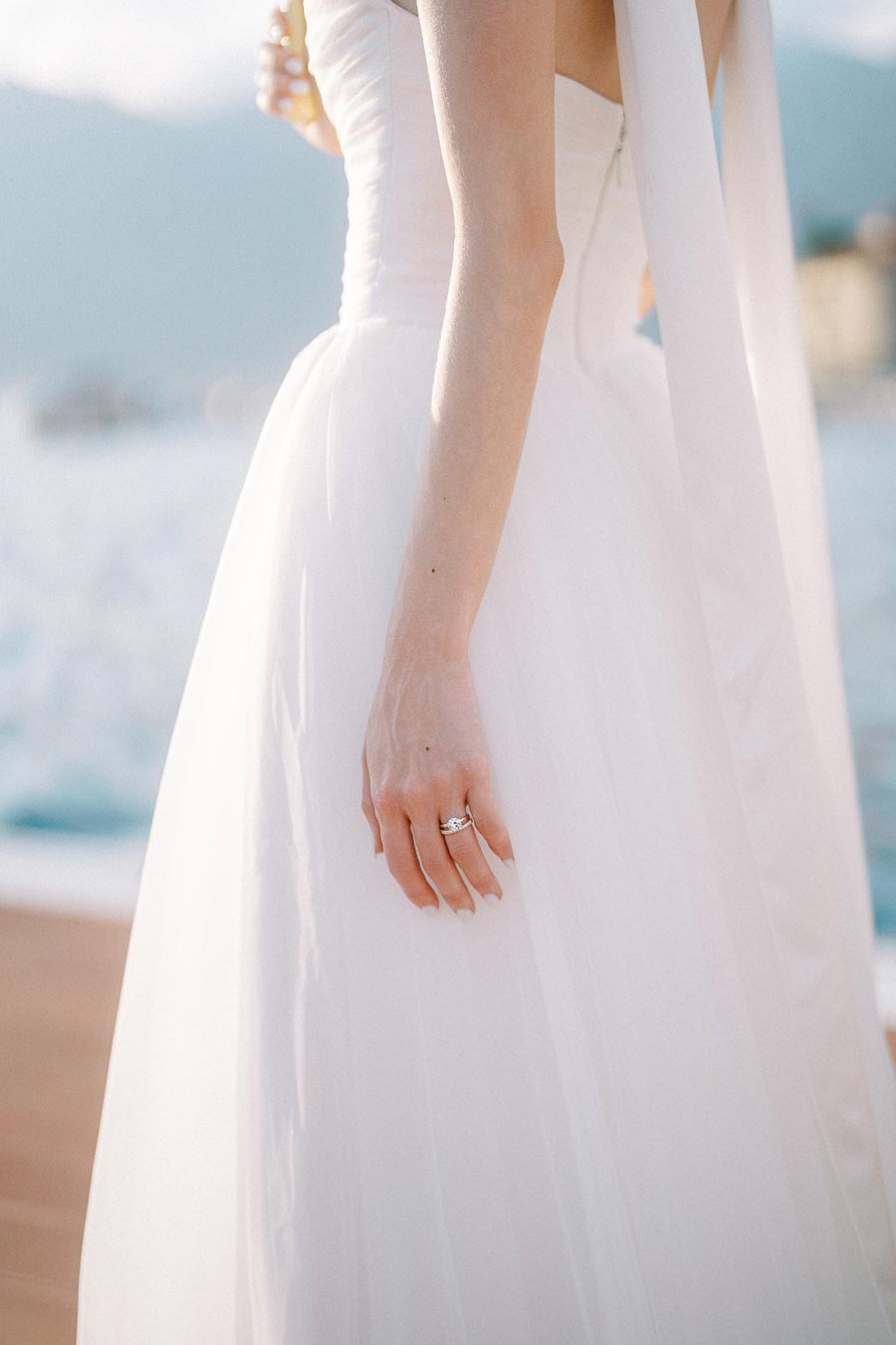 Elegant bride in a strapless white wedding dress with a sparkling diamond ring, standing by a scenic waterfront view.
