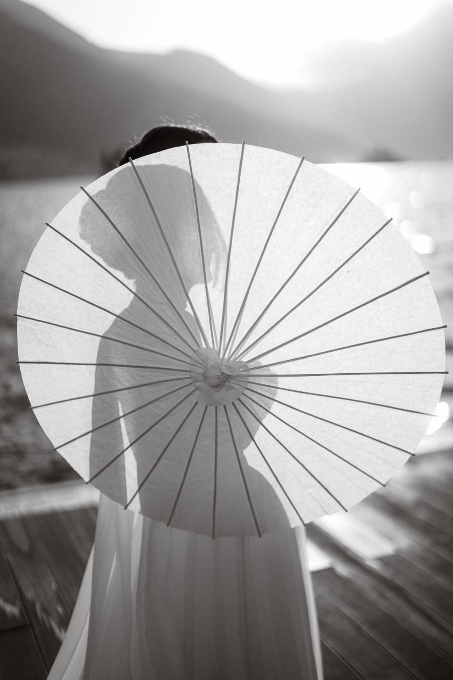 Black and white image of a woman holding a parasol in front of her face, with her silhouette visible through the fabric. The serene background features a lake and mountains, creating a romantic and mysterious atmosphere.