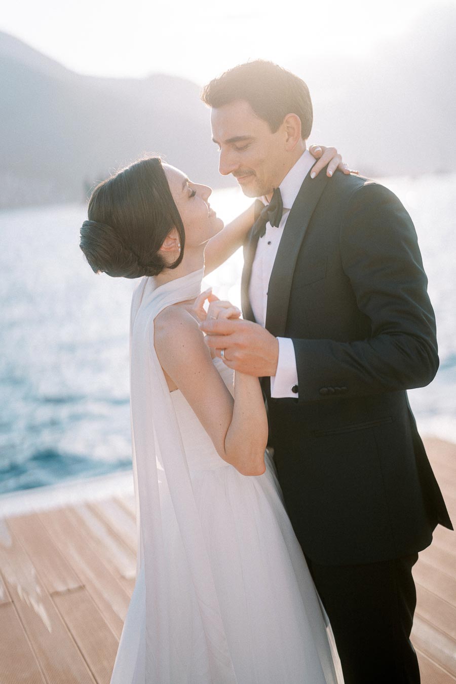 A couple dancing on a waterfront deck, with a scenic mountain view and sunlight softly illuminating their embrace. The woman is in a white dress and the man in a black suit, exchanging a tender moment by the water.