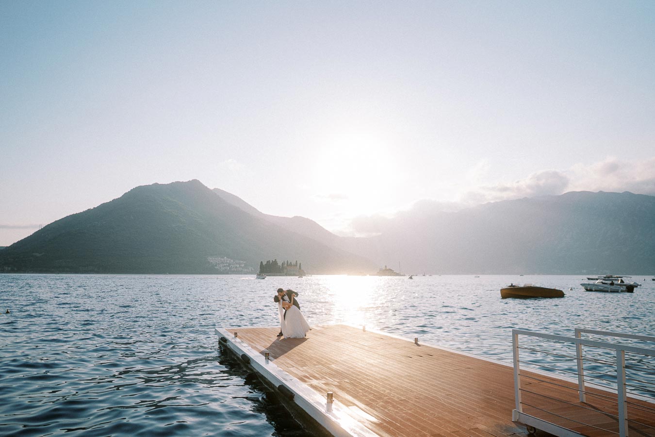 A newlywed couple kissing on a wooden dock at sunset with scenic mountains in the background and calm sea waters around.