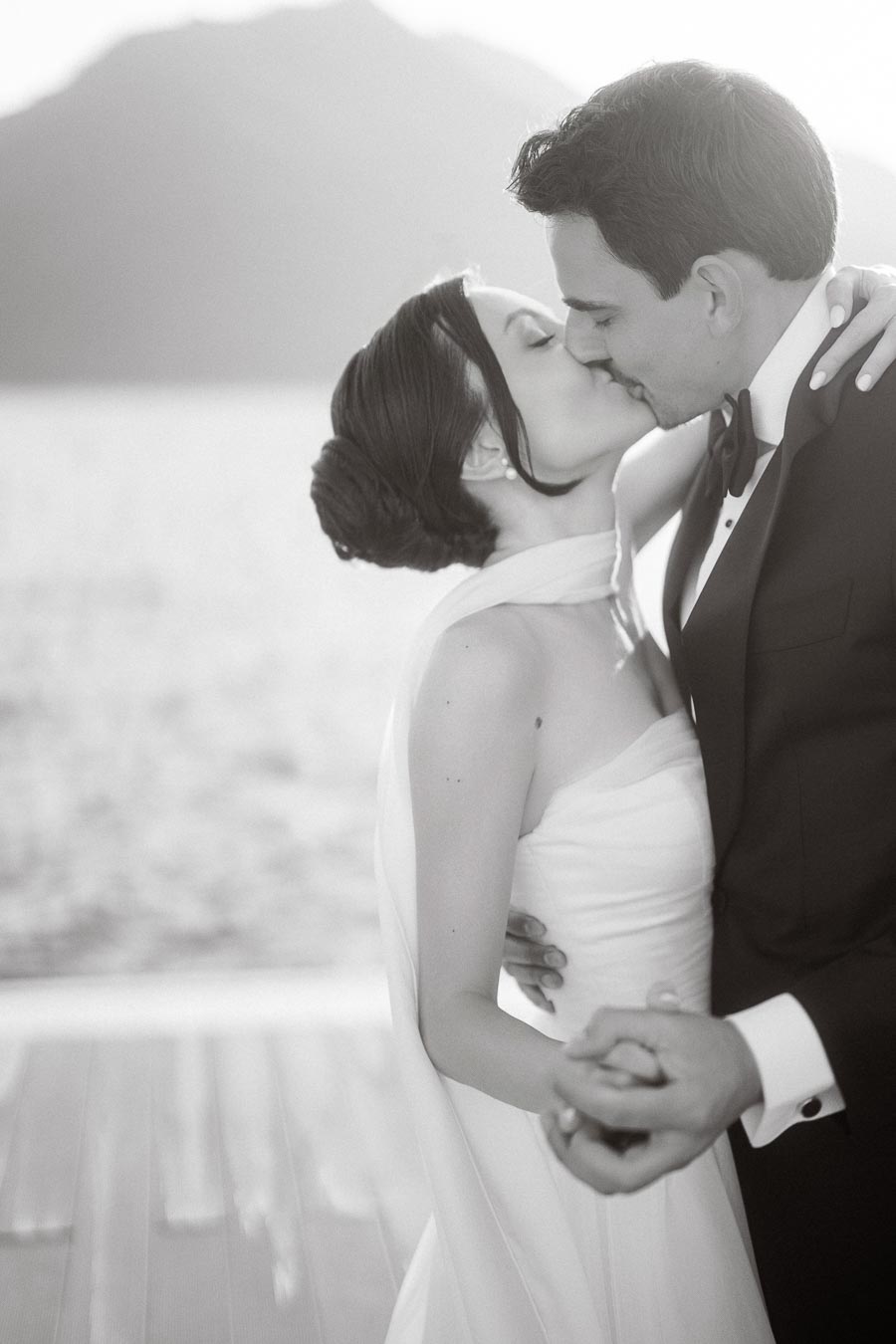 Bride and groom sharing a romantic kiss by the waterfront, dressed in elegant wedding attire with a scenic mountain backdrop.