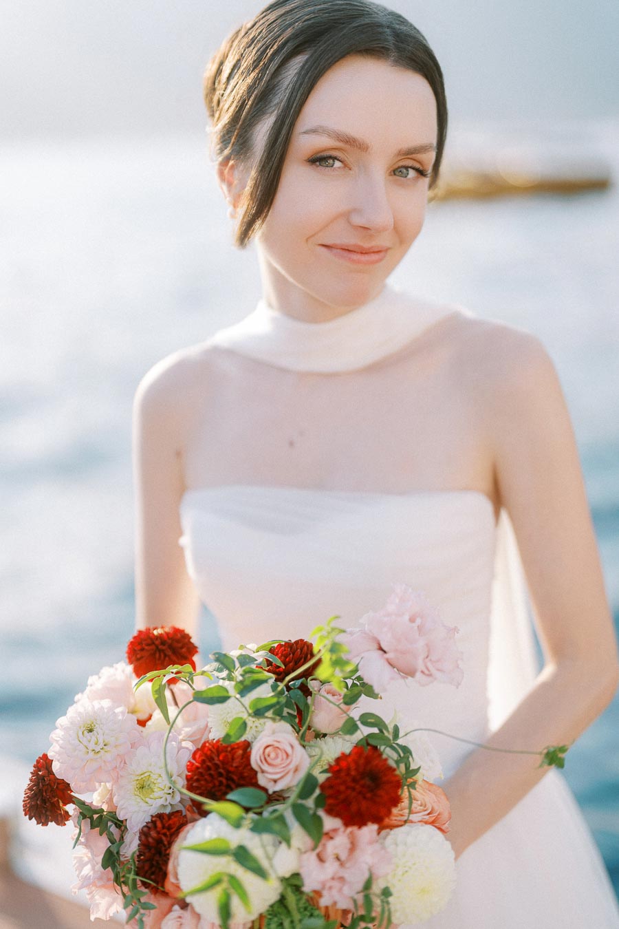 Young woman in a white wedding dress holding a bouquet of pink and red flowers, smiling by the sea on a sunny day.
