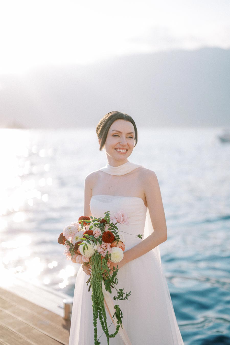 A smiling bride in a white strapless wedding dress holding a colorful bouquet stands on a wooden pier with a serene lake and mountains in the background, captured during golden hour.