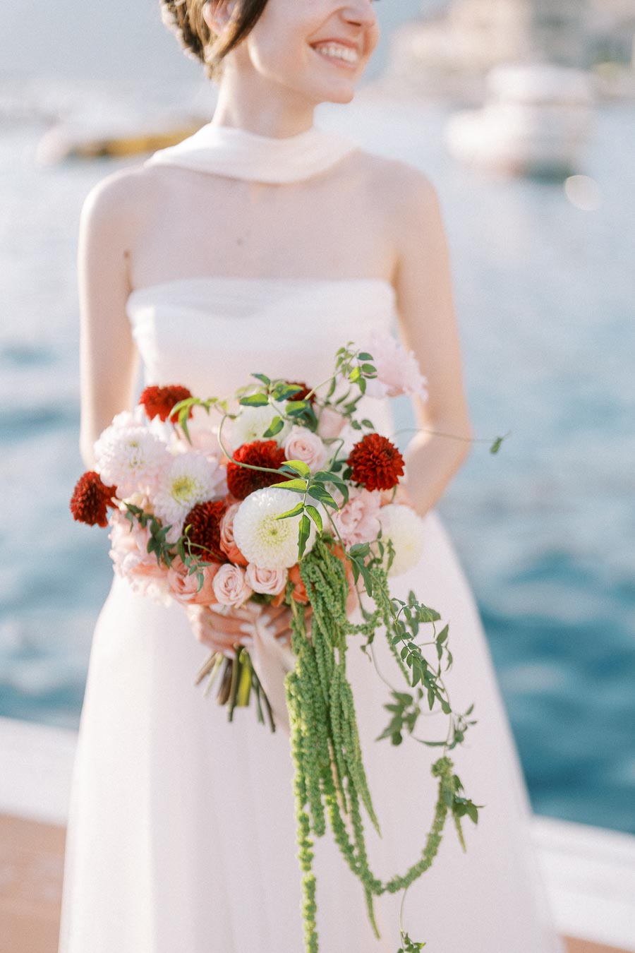 A bride in a strapless white wedding dress holding a vibrant bouquet of white, pink, and red flowers with greenery, standing in front of a blurred waterfront background.