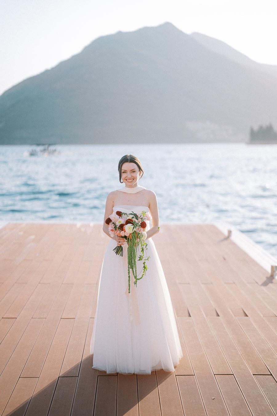 Beautiful bride in an elegant white wedding dress holding a bouquet of flowers on a wooden dock with a scenic mountain and ocean backdrop