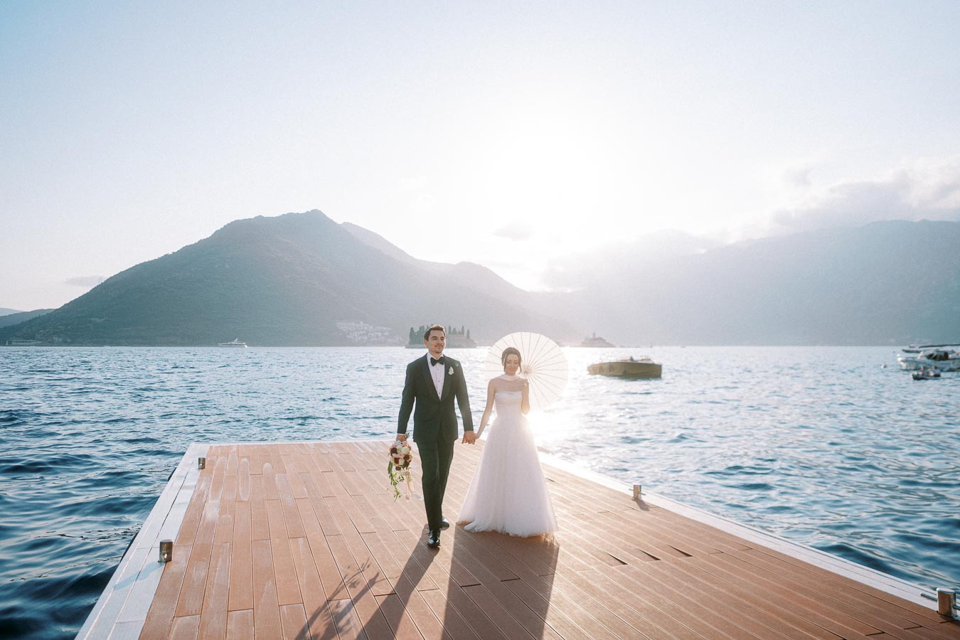 Bride and groom holding hands on a dock with mountains and water in the background, under a bright sky.