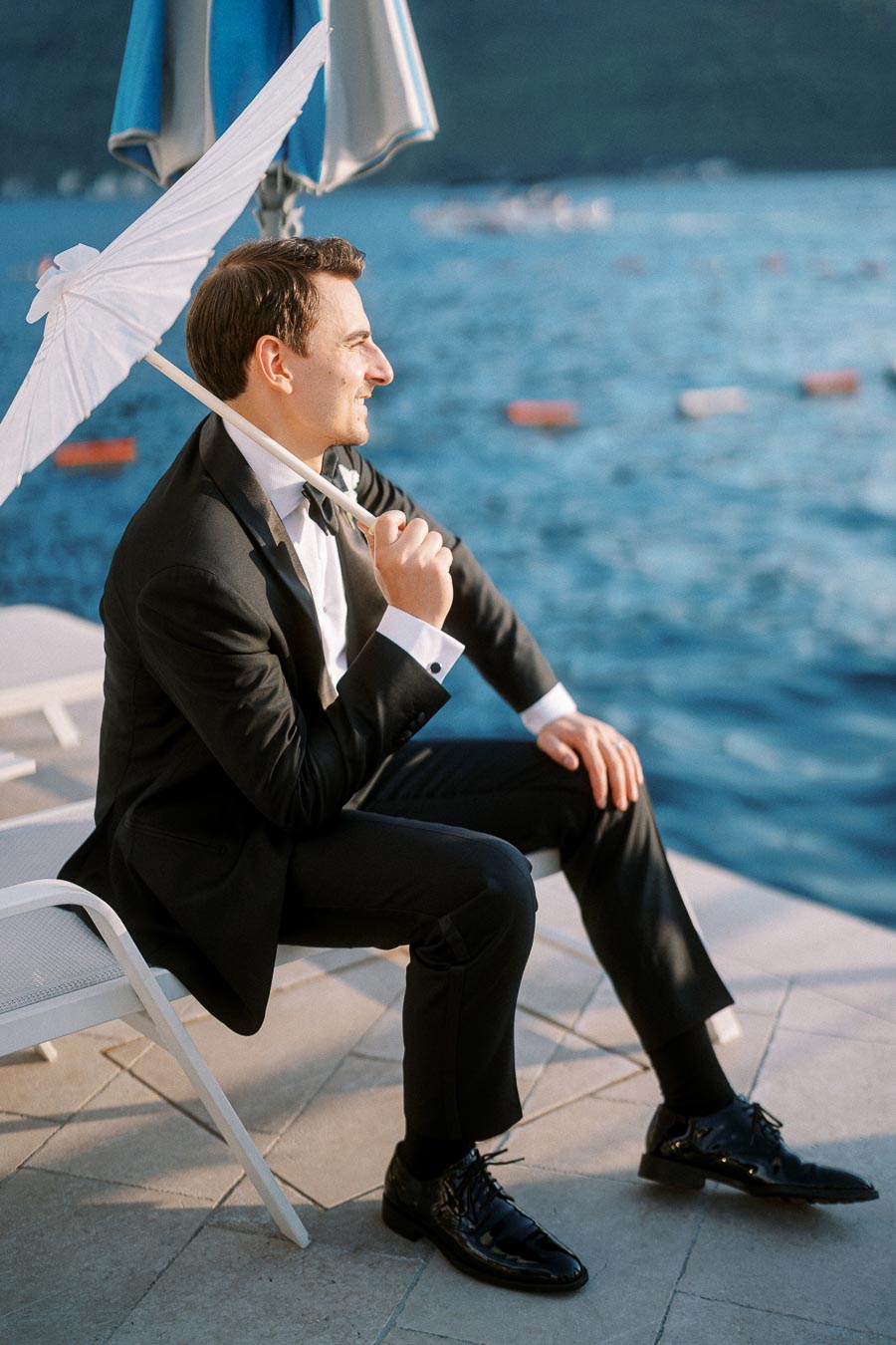 A man in a black suit sits by the waterfront holding a white umbrella, with a backdrop of blue water and distant boats under afternoon sunlight.