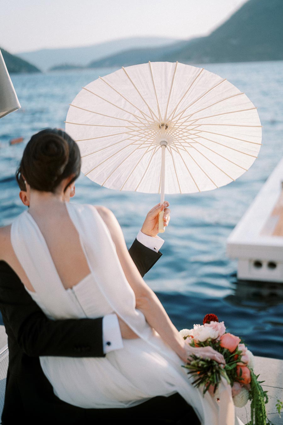 Romantic couple embracing by the ocean with a white parasol and a bouquet of flowers, capturing an elegant and serene wedding moment by the water.