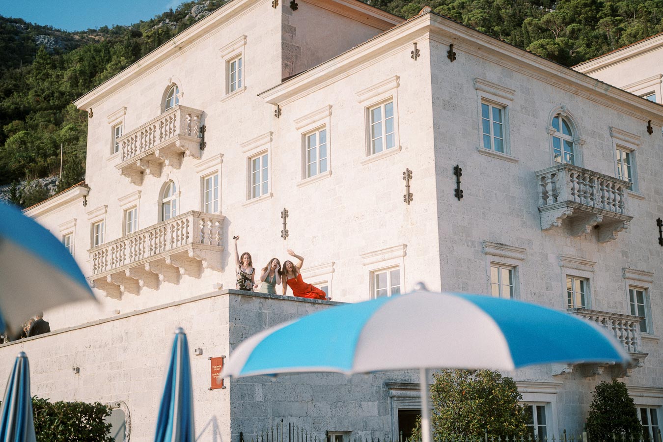 Three women on a balcony of a historic stone building, surrounded by lush greenery, with blue and white umbrellas in the foreground.