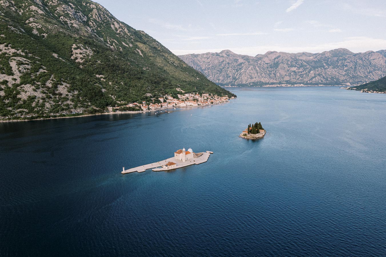 Aerial view of the Bay of Kotor in Montenegro featuring two small islands: Our Lady of the Rocks with its distinctive church and Saint George Island with lush greenery, set against a backdrop of rugged mountains and coastal town.