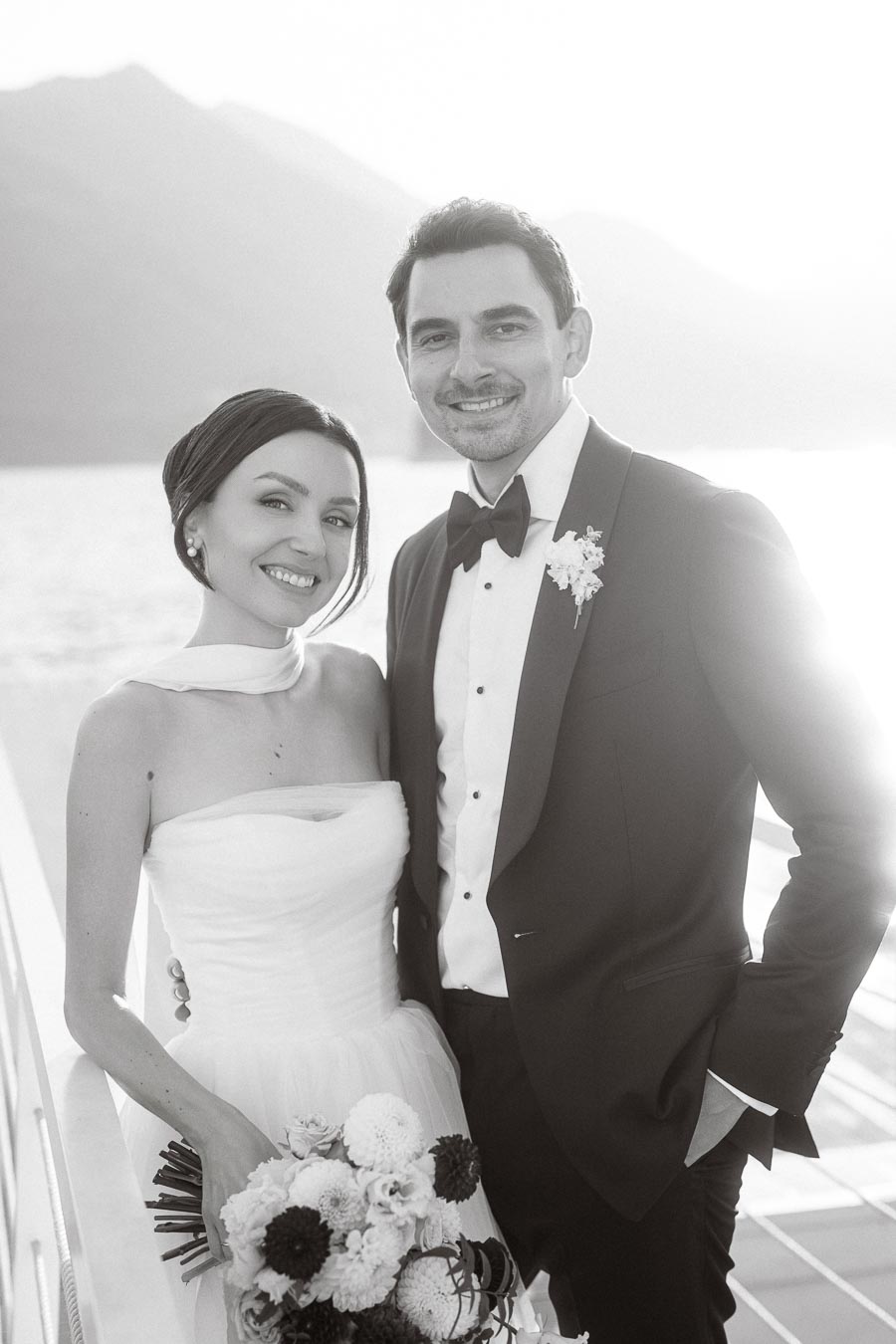 A couple celebrating their wedding, with the bride in an elegant strapless gown holding a bouquet of flowers and the groom in a classic tuxedo, smiling against a scenic lake and mountain backdrop in black and white.