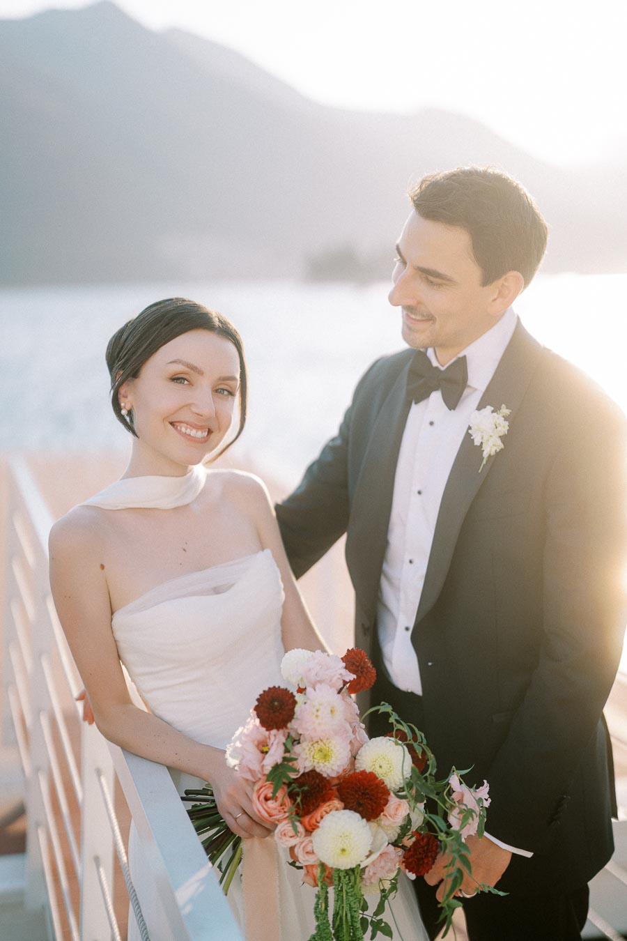 A happy bride and groom in elegant wedding attire, standing on a sunlit deck overlooking a serene body of water, with the bride holding a vibrant bouquet of flowers.