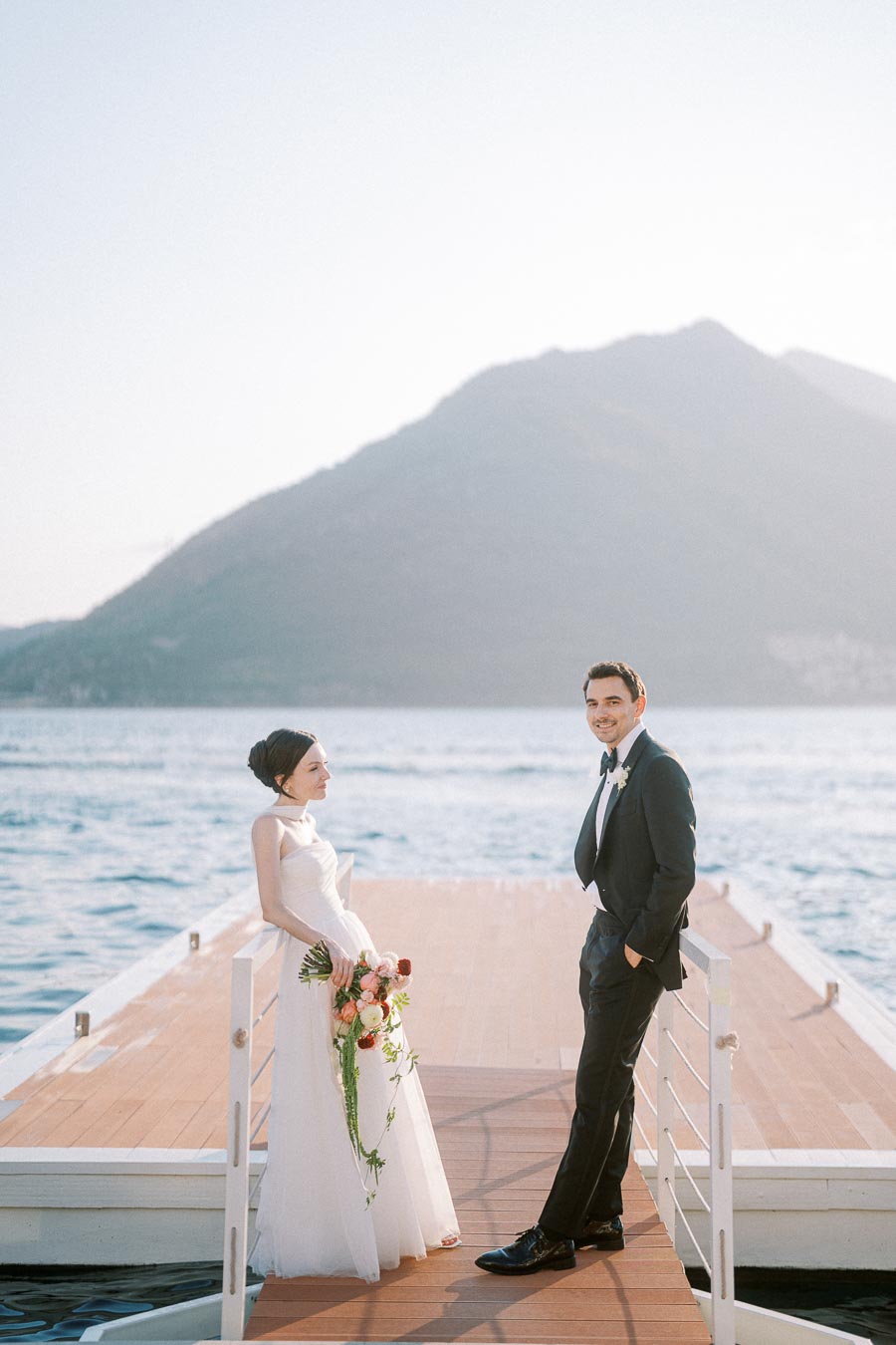 Elegant bride and groom posing on a wooden dock with a scenic mountain and lake backdrop, capturing a romantic wedding moment.