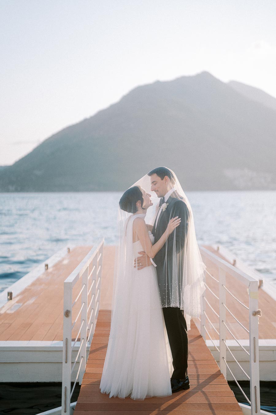 A bride and groom embrace on a wooden dock overlooking a serene body of water, with mountains in the background and sunlight illuminating their wedding attire.
