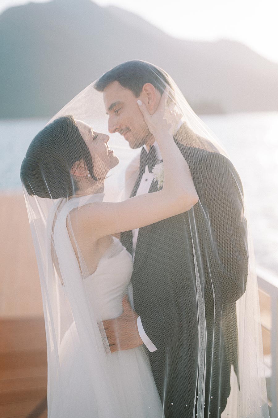 Romantic bride and groom embracing under a veil by a scenic waterfront at sunset, capturing a serene and intimate wedding moment.