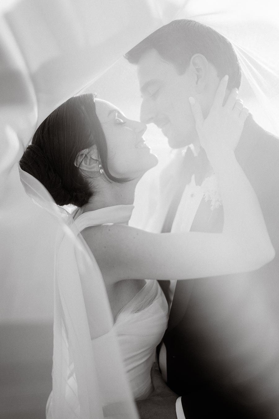 Black and white photo of a bride and groom embracing under a veil, capturing a romantic and intimate wedding moment.