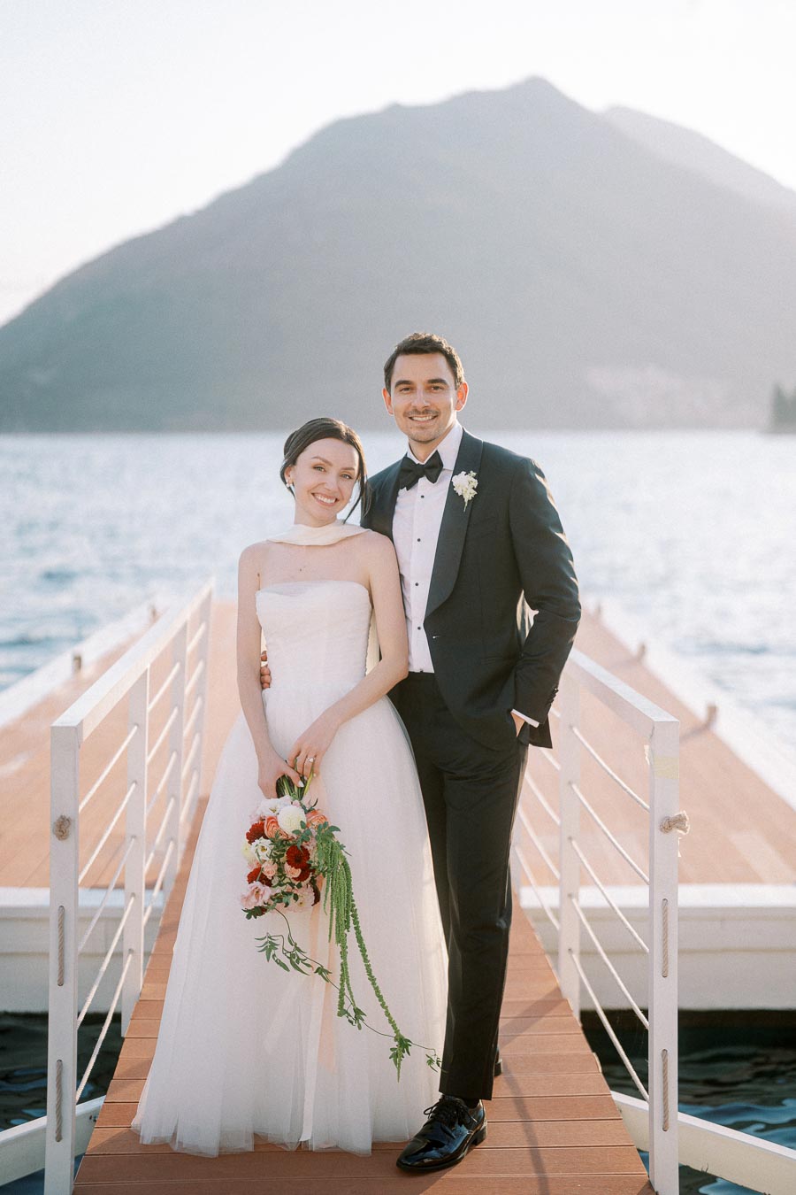 Happy couple in wedding attire standing on a dock with a scenic mountain and lake backdrop, bride holding a floral bouquet.