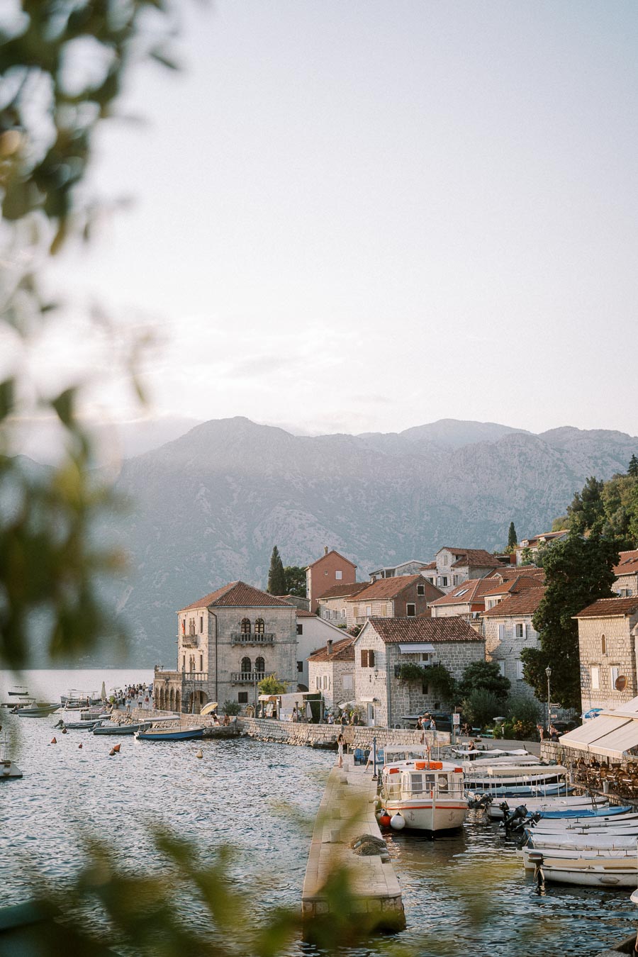 Scenic view of a Mediterranean coastal village with stone buildings and red-tiled roofs by the sea, boats docked along the waterfront, and mountains in the background.