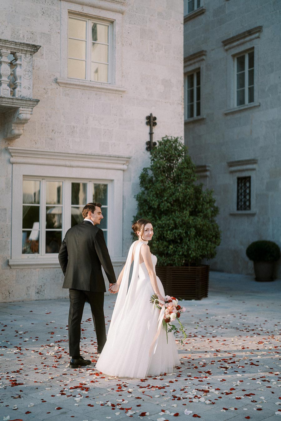 A newlywed couple in elegant wedding attire walking hand in hand on a flower-petal-strewn pavement in front of a historic stone building, surrounded by greenery.