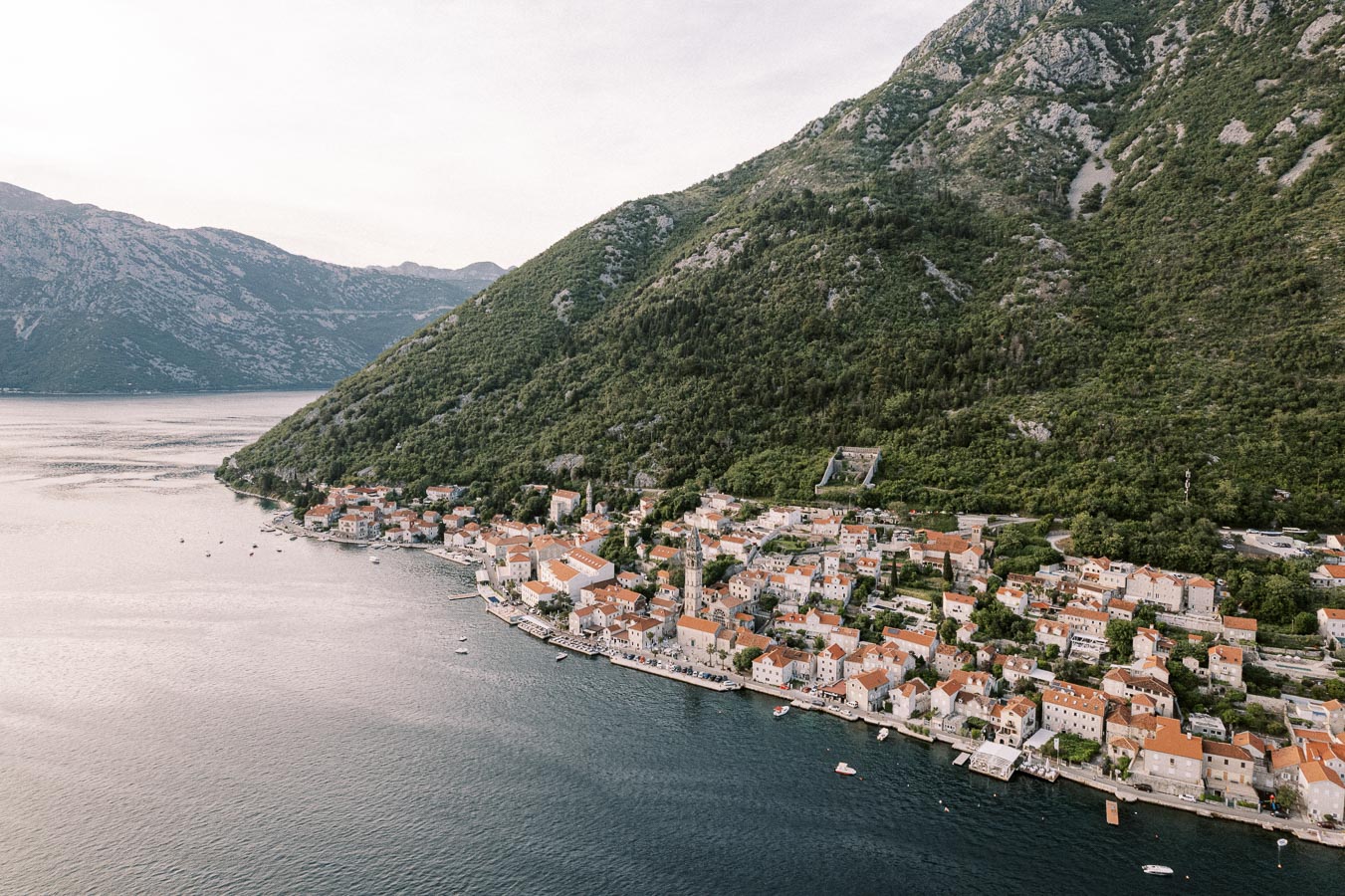 Aerial view of a picturesque coastal town with terracotta rooftops nestled against a lush green mountain, adjacent to serene blue waters.