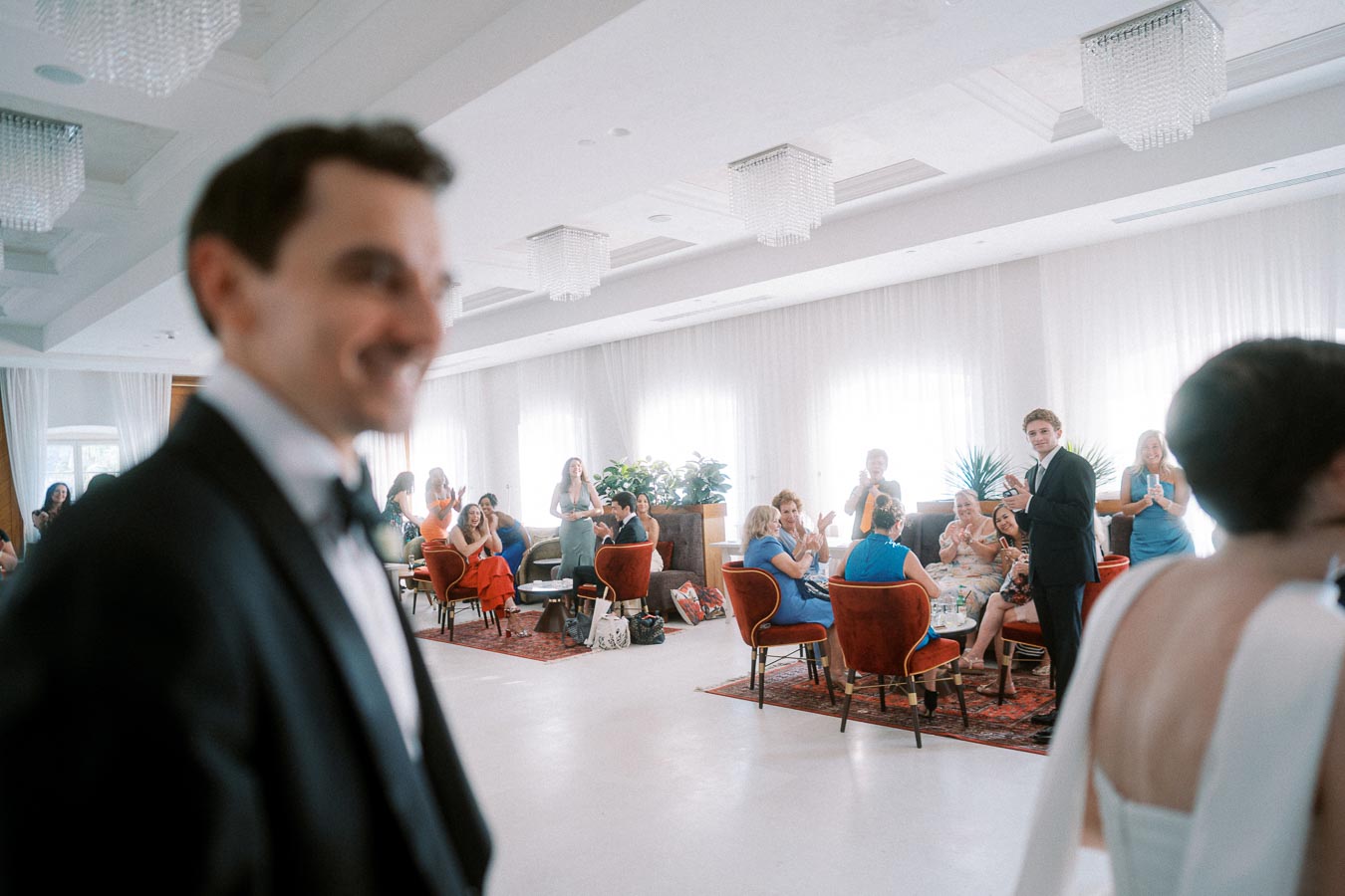 A joyful wedding reception in a bright, elegant room with guests applauding and taking photos, featuring a smiling groom in the foreground and a bride in a white dress.