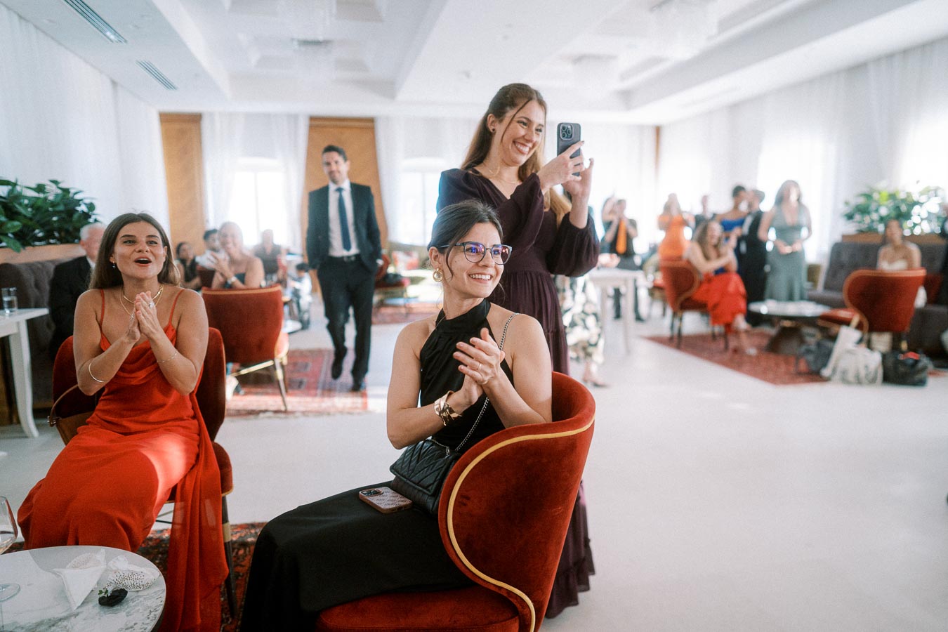 Women in elegant dresses happily clapping and taking photos during a formal indoor event, with other attendees in the background.