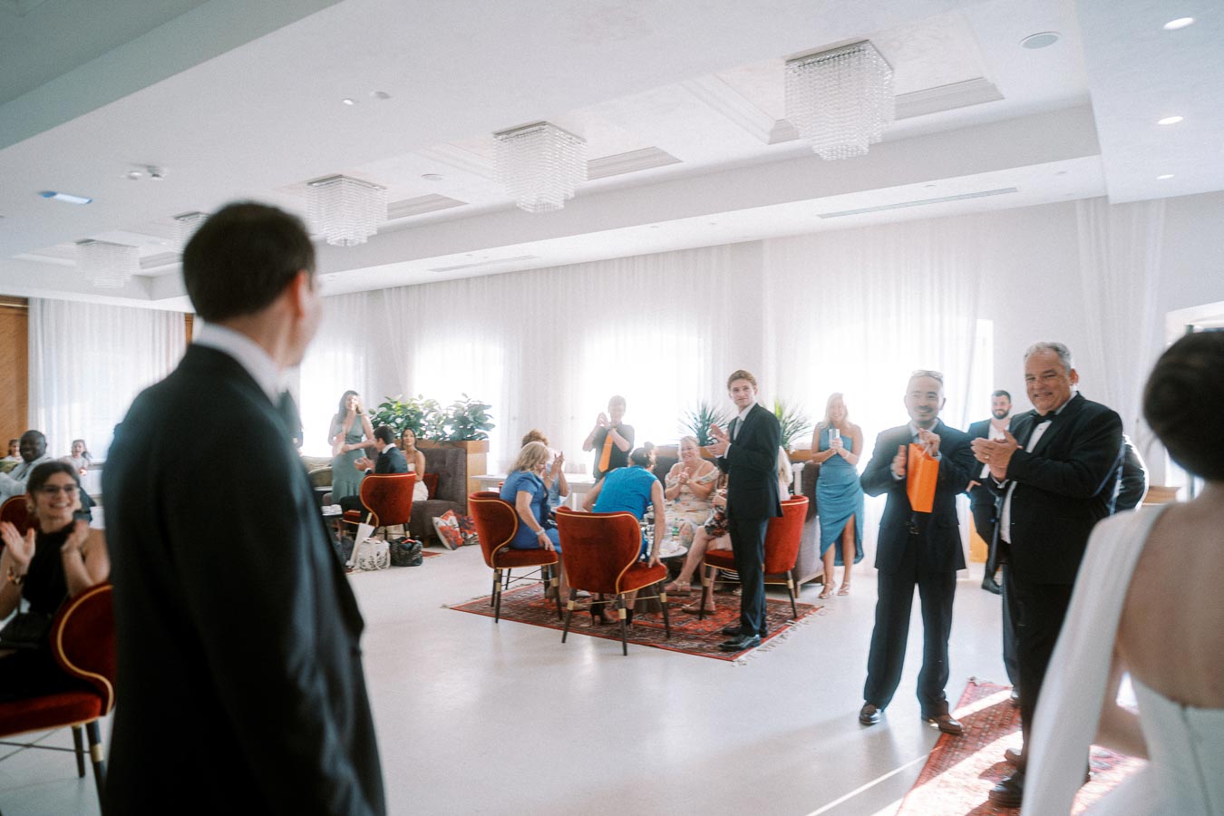 Formal gathering in a bright, elegantly decorated room with attendees wearing suits and dresses. People are seated around tables and standing, clapping and smiling, indicating a celebratory or social event. The room features large windows with natural light and chic furnishings.