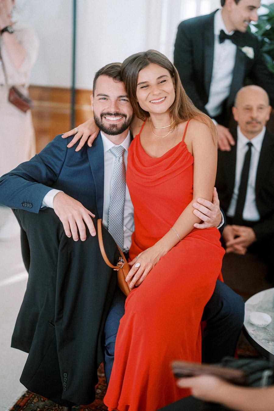 A woman in a red dress smiling while sitting on the lap of a man in a blue suit at a formal event, surrounded by other guests in evening attire.
