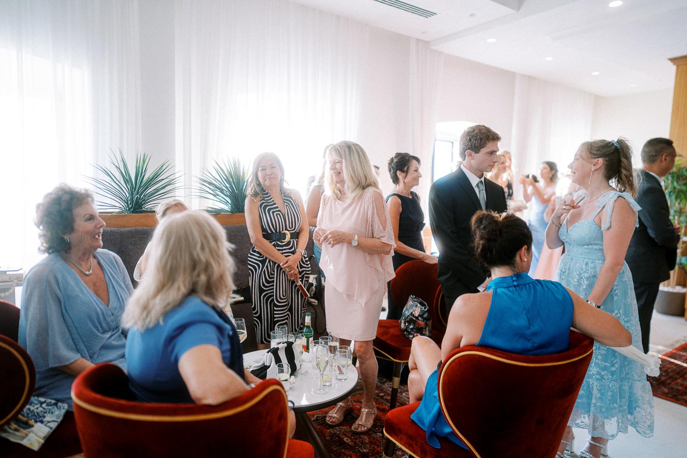 Group of elegantly dressed people socializing at an indoor gathering or celebration, enjoying conversations and drinks in a warmly lit room with stylish decor.