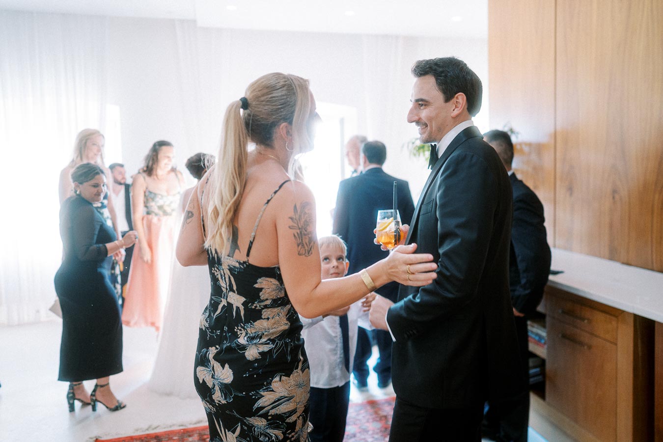 A group of elegantly dressed people, including a woman in a floral dress and a man in a suit holding a cocktail, engage in lively conversation at a wedding reception.