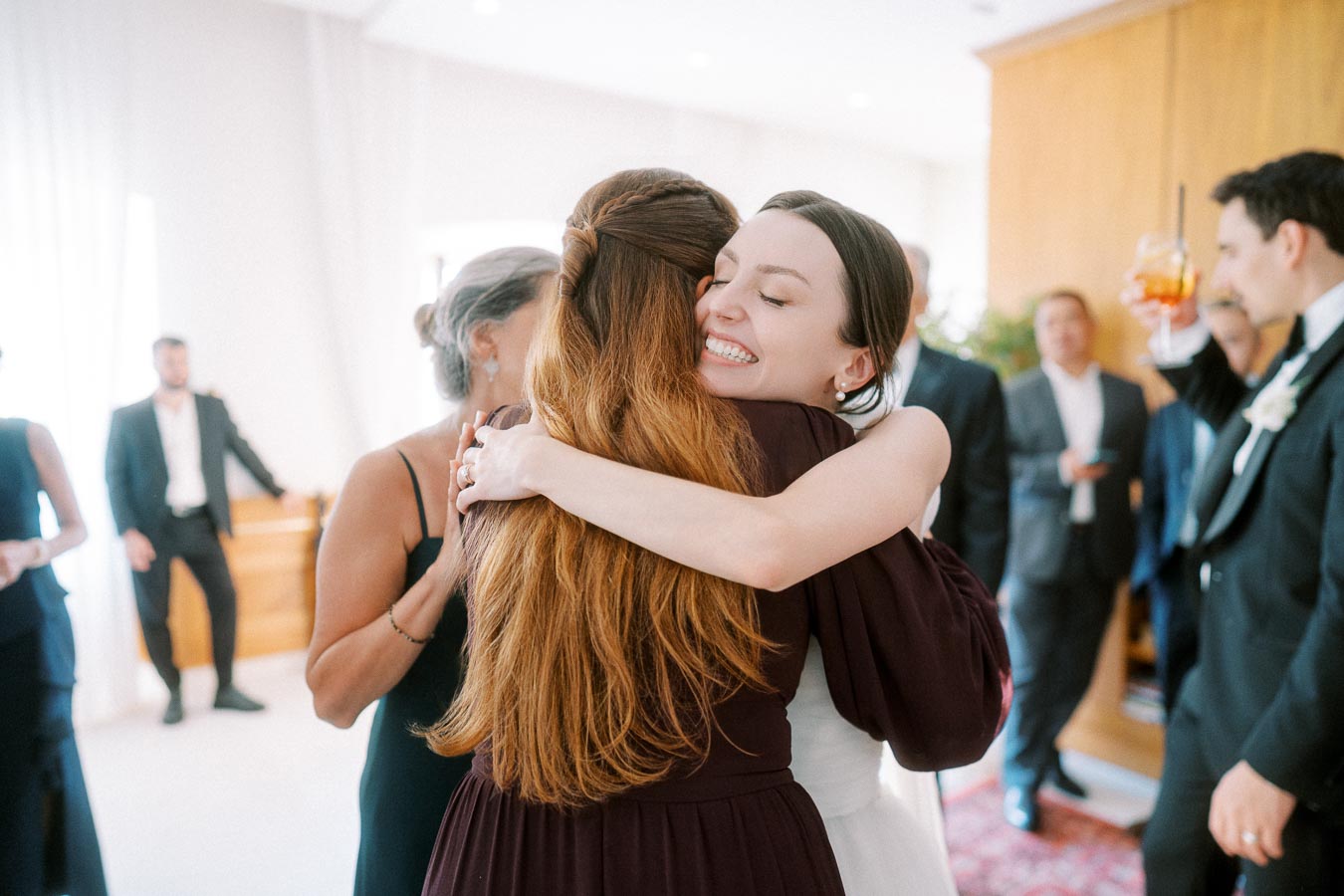 A joyful woman with dark hair warmly embraces a friend in a crowded, elegantly decorated gathering, surrounded by people dressed in formal attire, capturing a moment of celebration and connection.