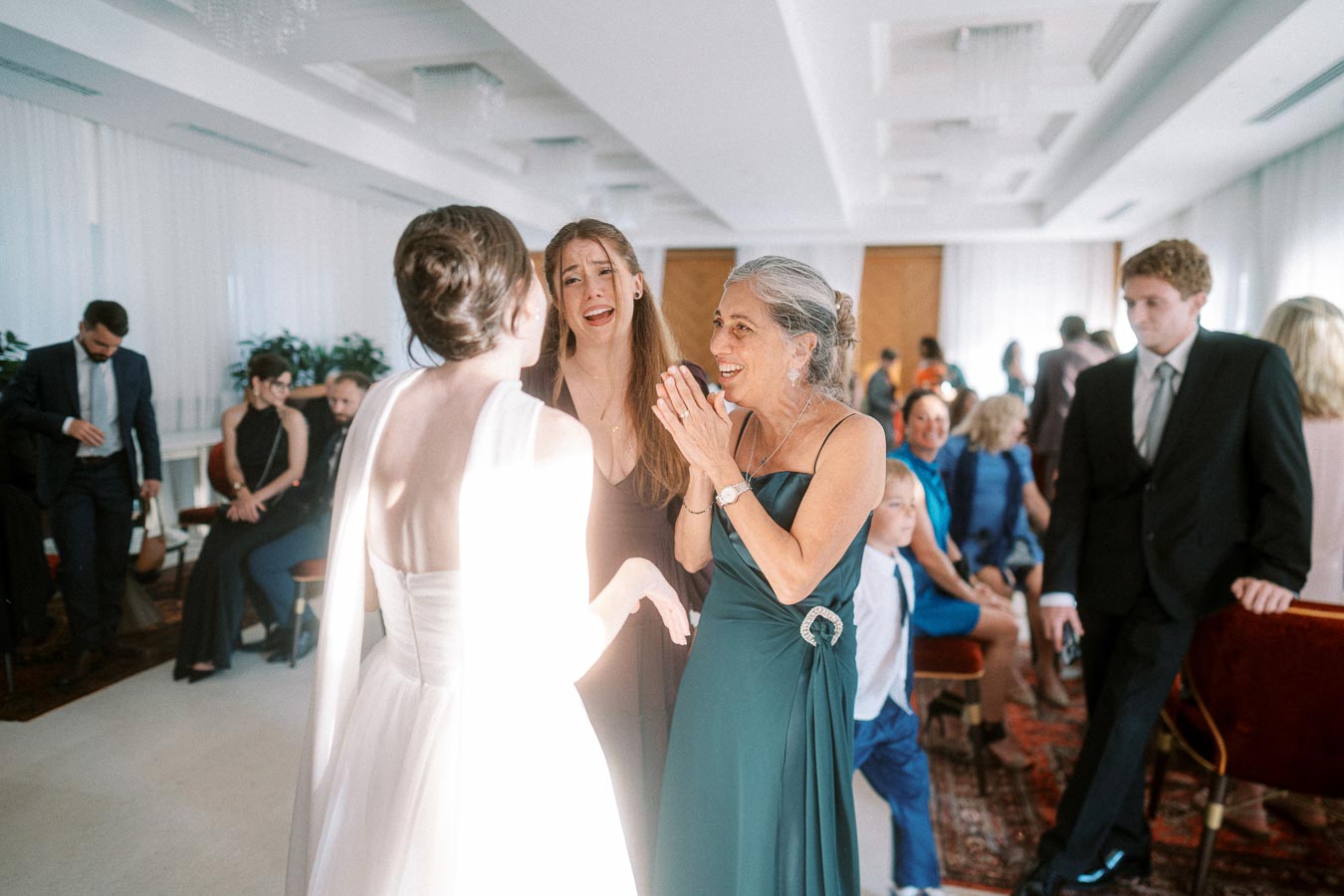 A bride in a white gown interacting joyfully with two women, one in a dark dress and the other in an emerald green gown, surrounded by wedding guests in a bright, elegantly decorated venue.