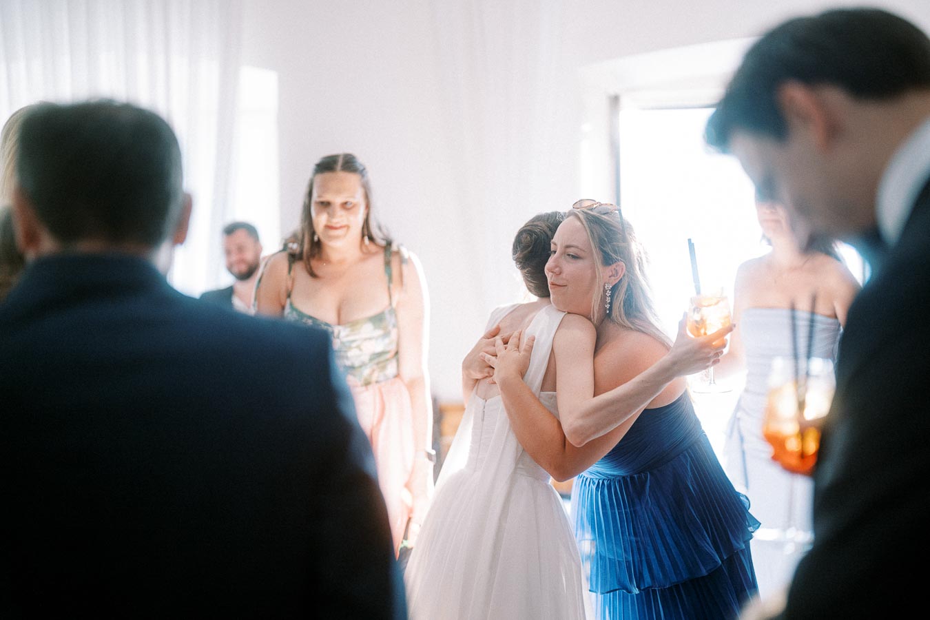 Wedding guests at a reception, with two women embracing warmly, one in a white dress, the other in a blue dress, surrounded by people in formal attire holding cocktails.