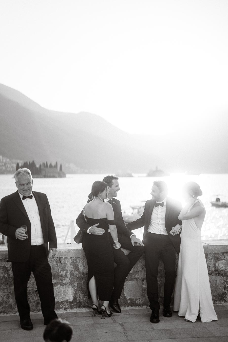 Black and white photo of elegantly dressed people at a waterfront event during sunset, with mountains and a calm sea in the background.