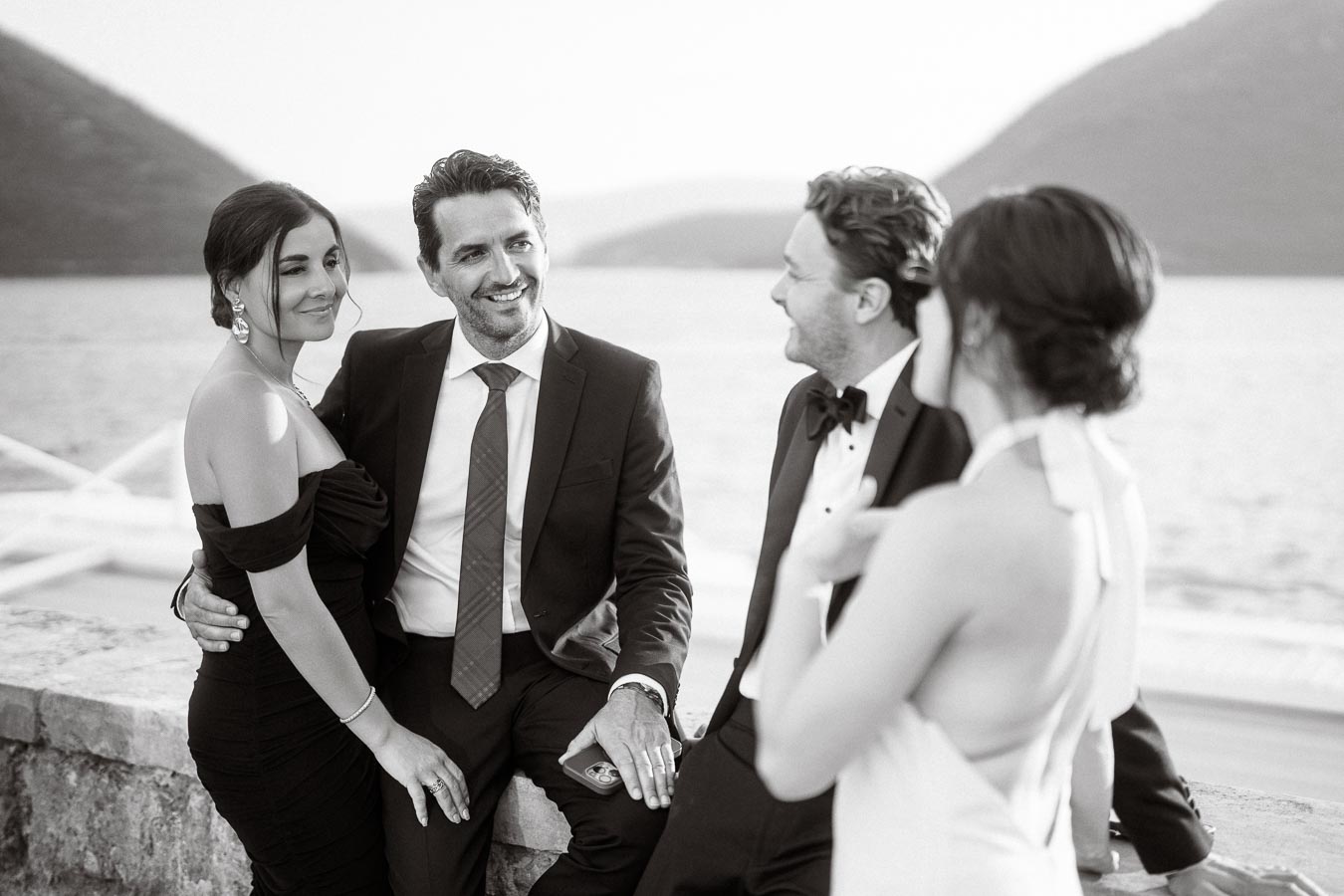 A group of elegantly dressed people socializing by the waterfront, with men in suits and women in formal gowns, set against a scenic backdrop of mountains and water.