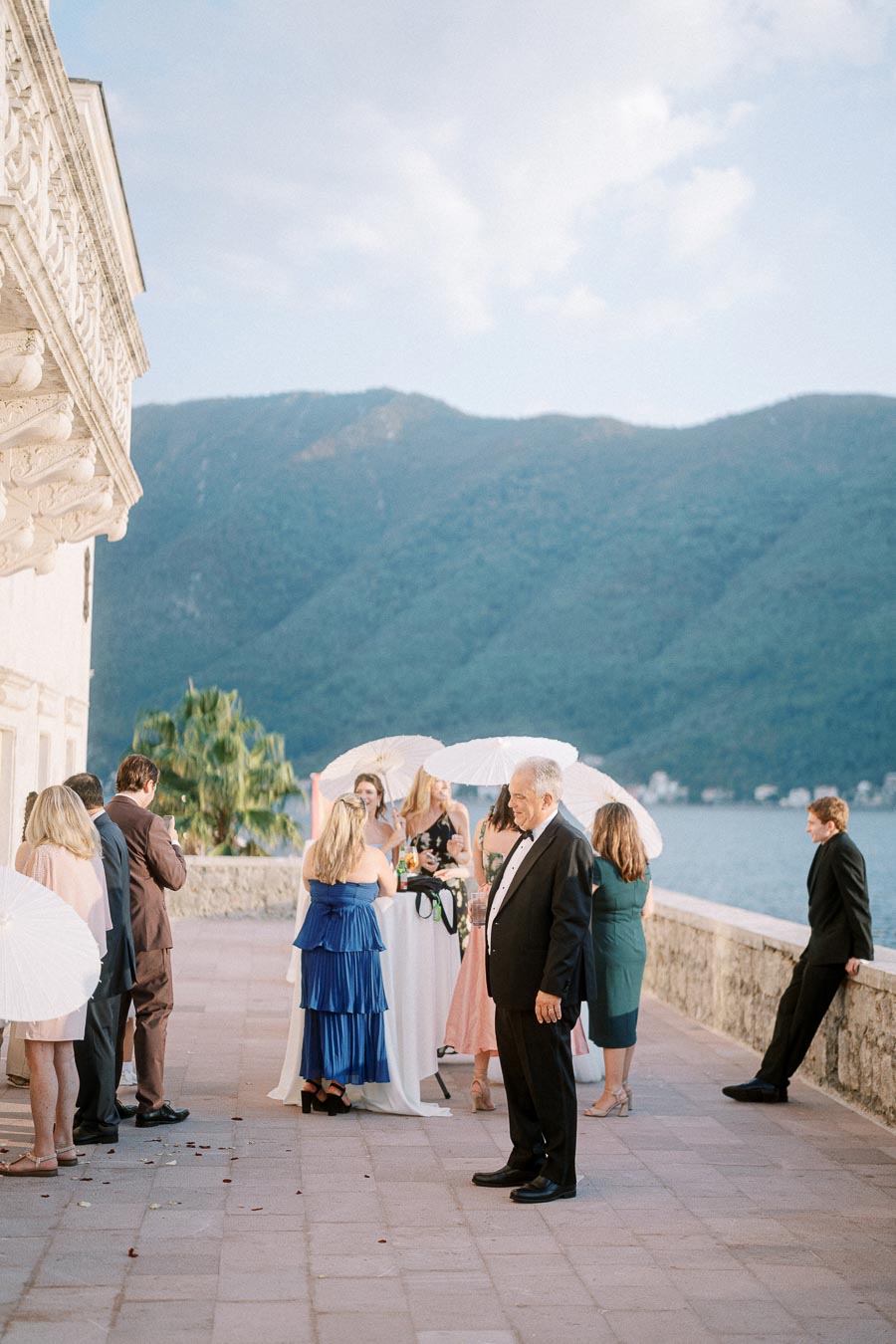 Elegant outdoor wedding reception on a terrace overlooking mountains and a lake, guests in formal attire holding parasols, creating a picturesque backdrop.